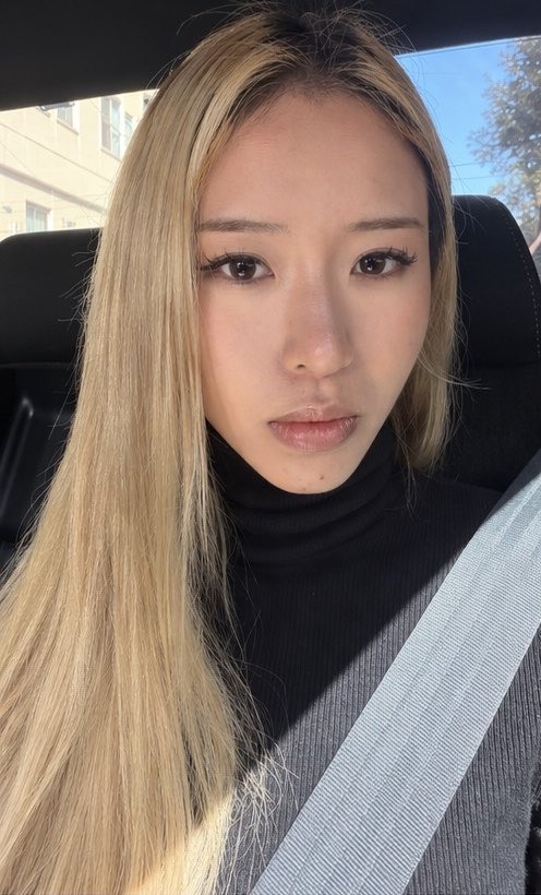 A woman photographed in a car, long straight blonde hair worn flat with a center part.